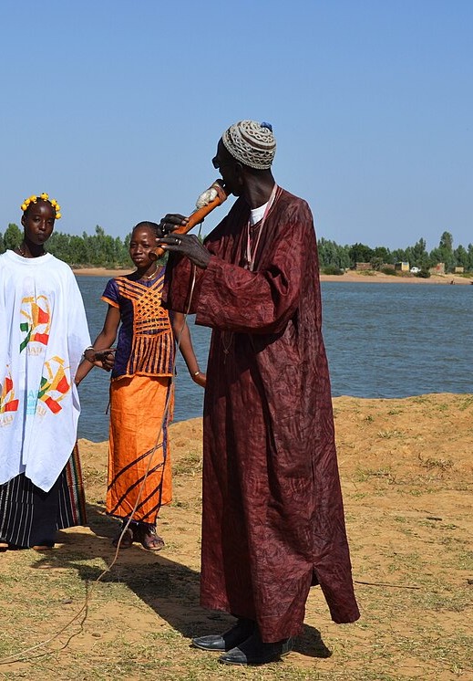 Children in white robes stand by a river while a man in a long robe plays a flute held at an angle