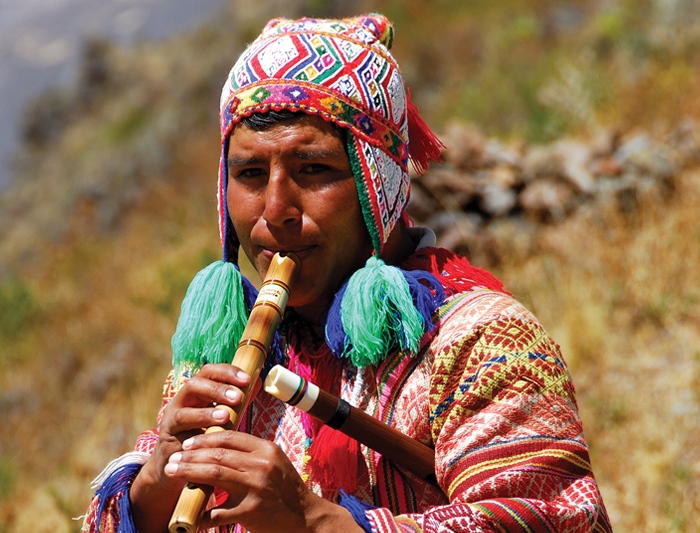 A Quechua musician in a bright woven hat and poncho plays a wooden quena flute outdoors in Peru