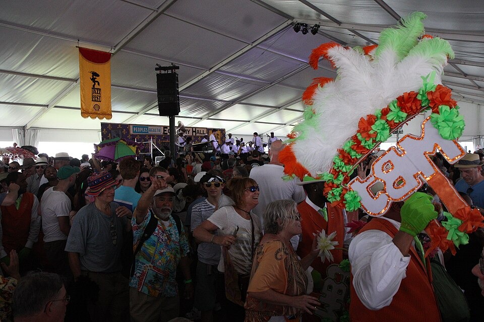 A crowd dancing under a festival tent while a dancer in a feathered costume holds up a decorated fan; a brass band plays on the stage behind