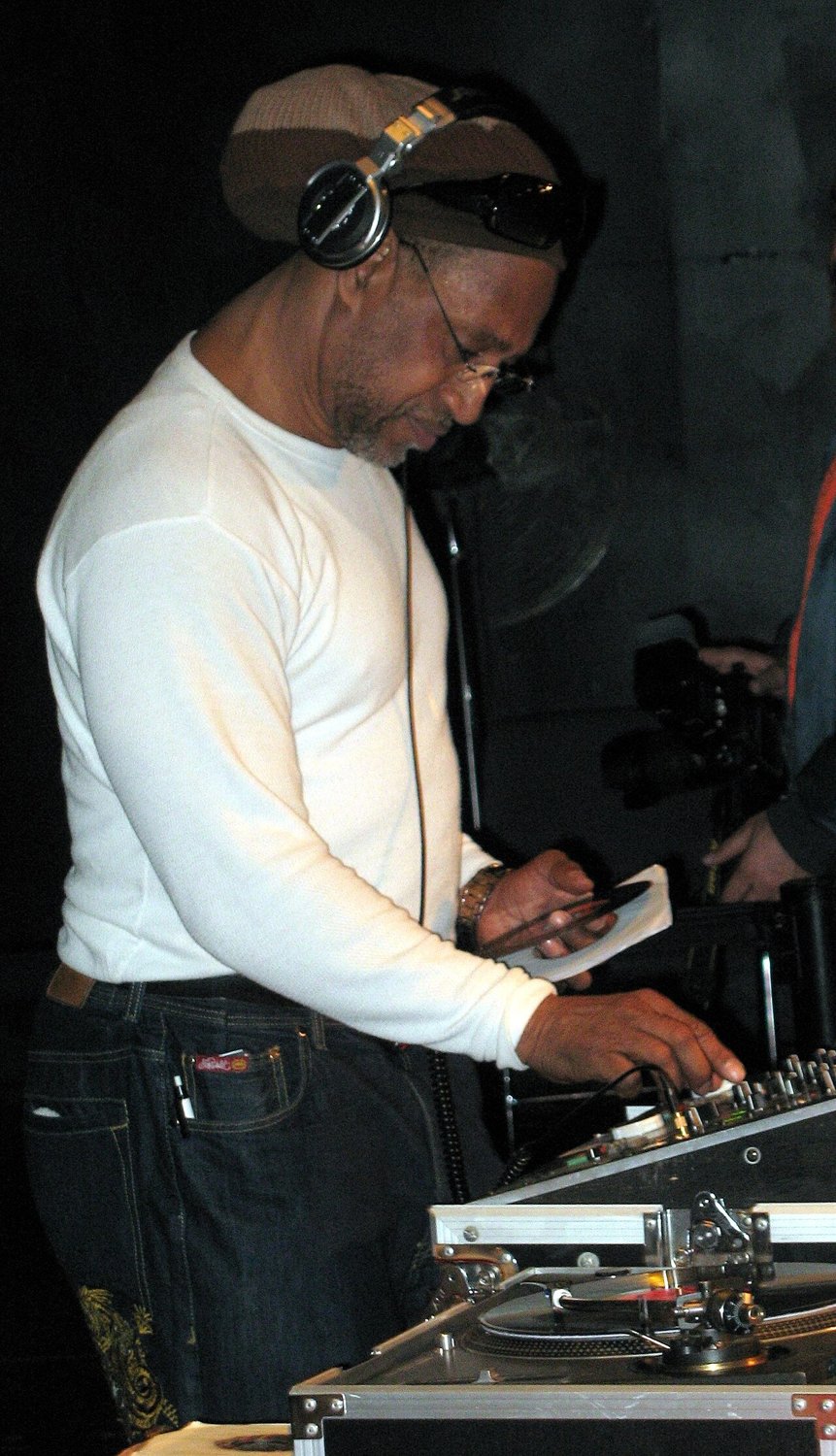 DJ Kool Herc in a white shirt and headphones, looking down at a record sleeve in one hand, his other hand on a DJ mixer beside a turntable