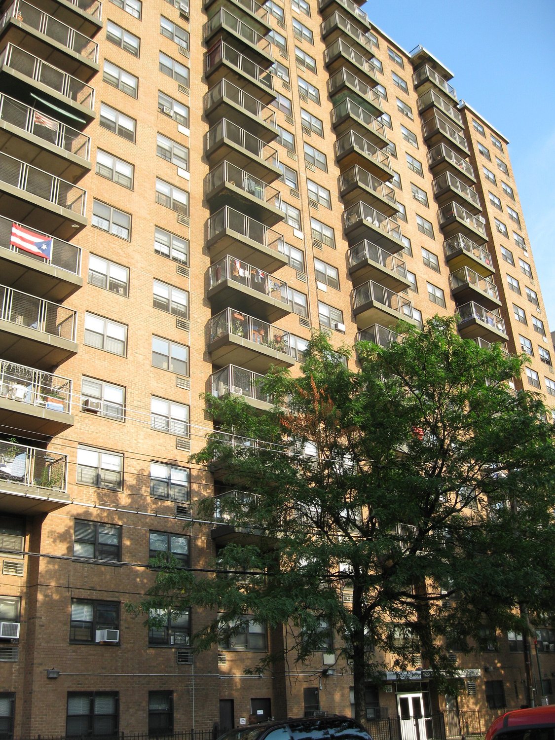 A tall brick apartment building with balconies, a Puerto Rican flag on one balcony and a tree in front