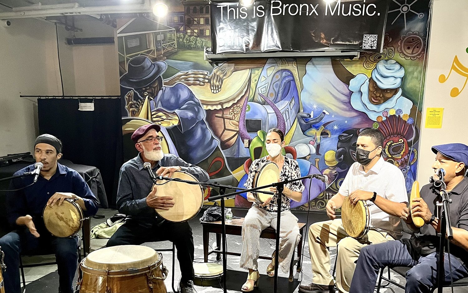 Five musicians seated in front of a colorful mural under a banner that reads This is Bronx Music: four play round hand drums called panderos, one plays a güiro, and a barrel drum stands in front