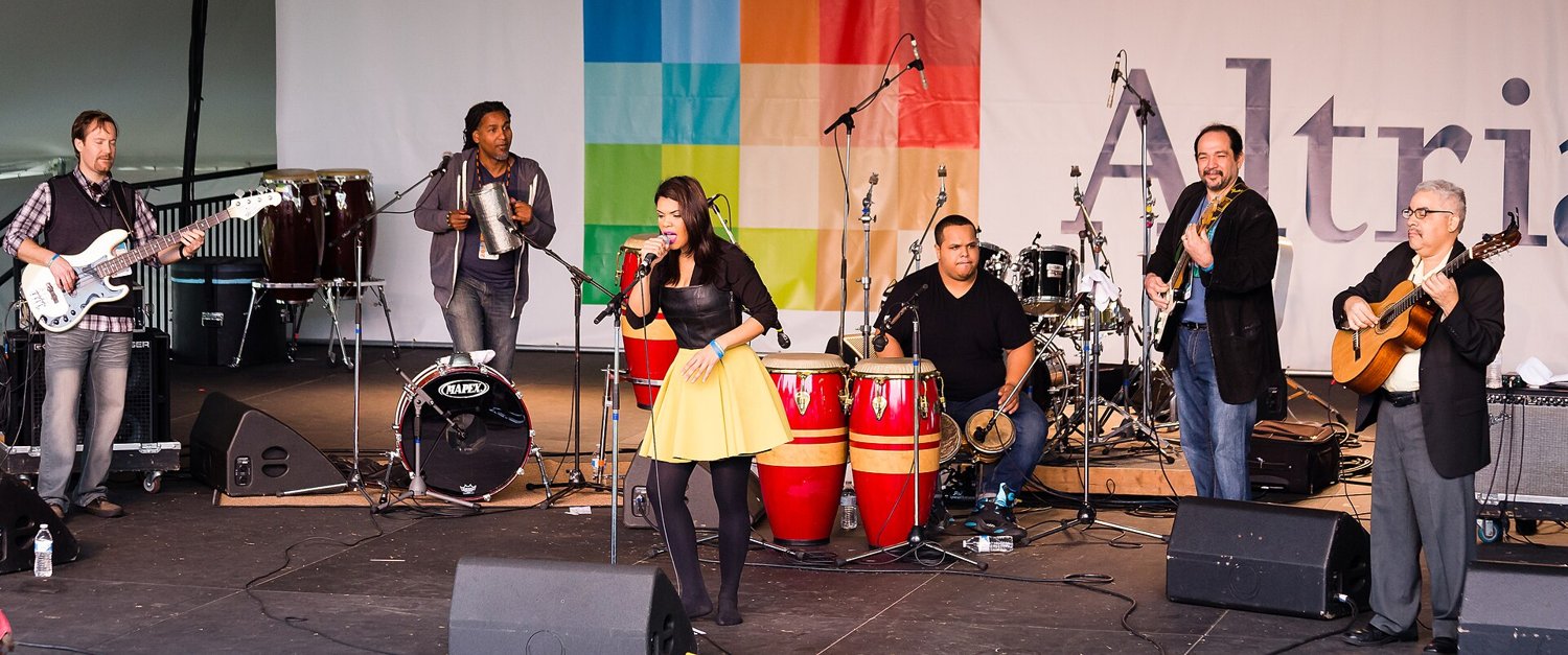 A bachata band on an outdoor stage: a bass player, a man scraping a metal güira, singer Andre Veloz in the center, a drummer at bongos and congas, and two guitarists
