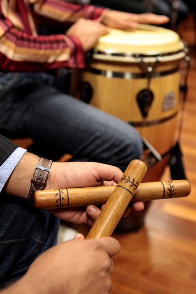 Two hands holding a pair of carved wooden claves crossed over each other, with a conga drum and its player behind them
