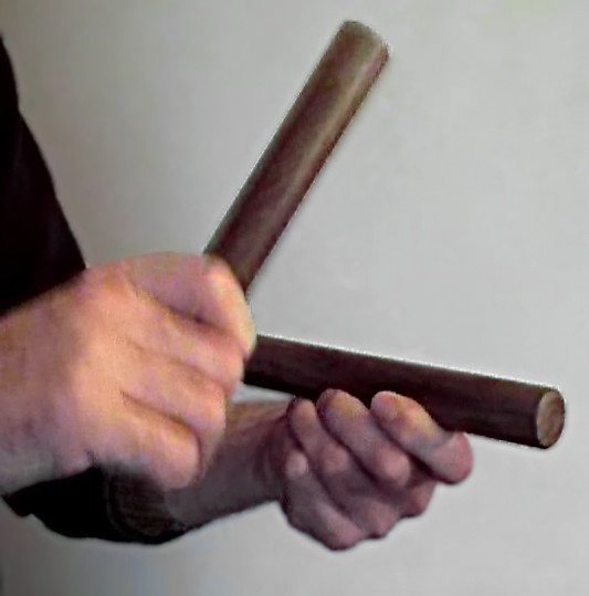 Two hands with claves: one wooden stick rests across the fingers of a cupped hand while the other hand holds the second stick to strike it