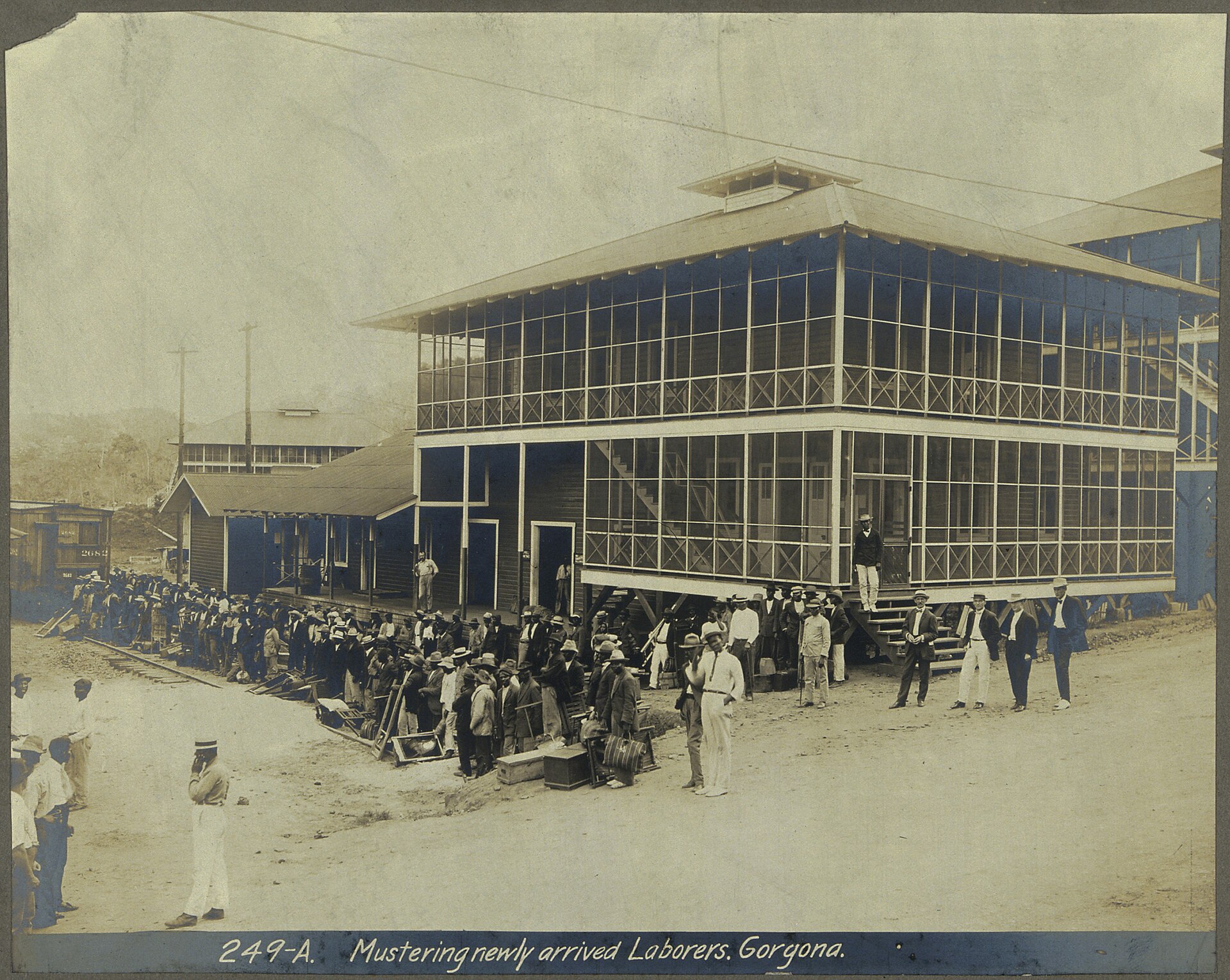 An old photograph of a long line of workers with hats and luggage beside a two-story screened building at a railway station