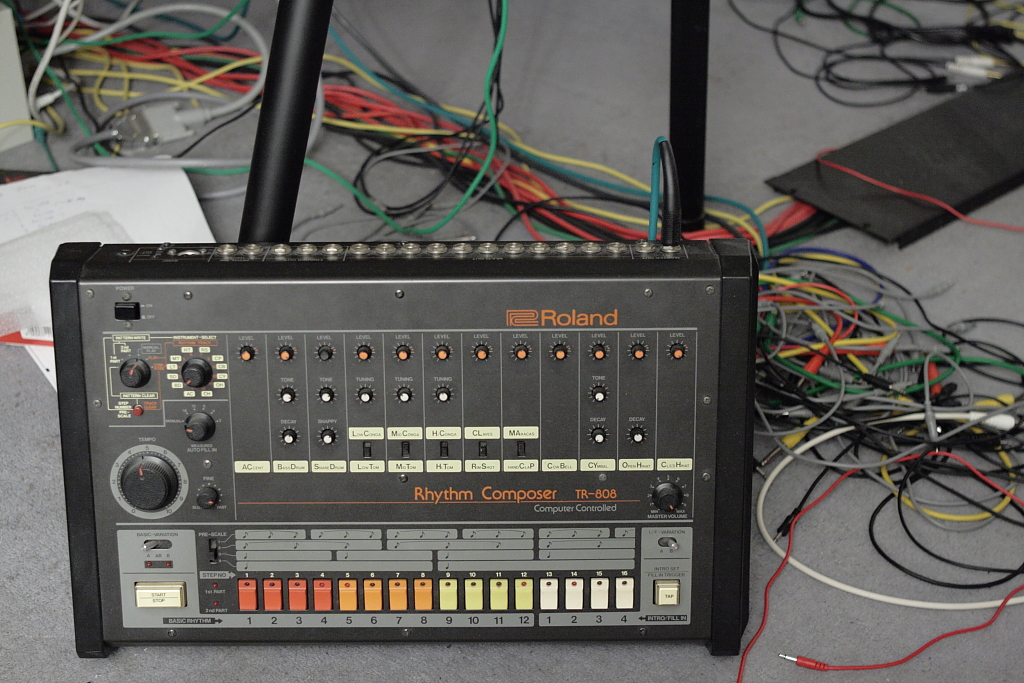A Roland TR-808 drum machine on a studio floor, with rows of orange knobs and sixteen colored step buttons