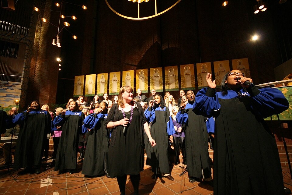 A soloist in a blue and black robe sings into a microphone with one hand raised while a gospel choir stands behind her